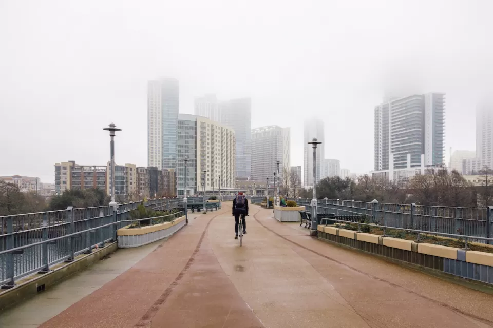 One cyclist rides on pedestrian bridge with foggy Austin, Texas skyline in background.